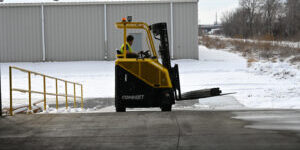 Operator driving a yellow CombiLift forklift outdoors on a snowy loading area near an industrial building.